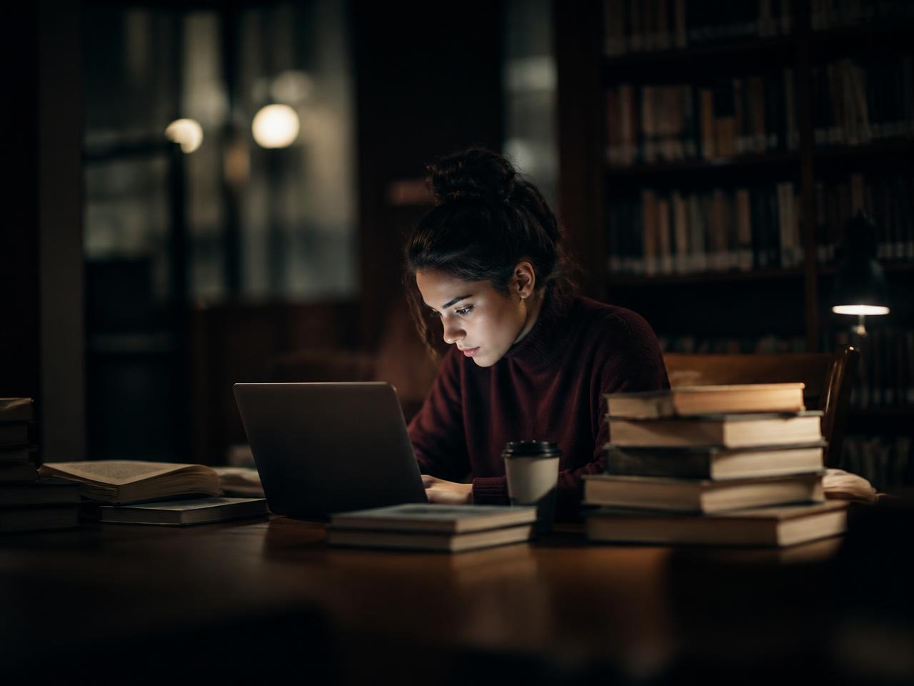 Une étudiante travaille seule le soir avec son ordinateur et des livres à la bibliothèque